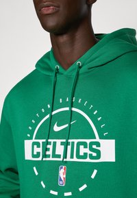Személy zöld Boston Celtics kapucnis pulóvert visel, amelyen látható a Nike logó és az NBA embléma a mellkason.