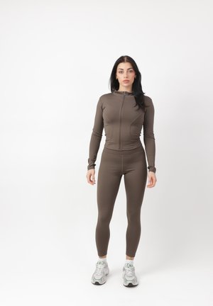 Kvinde stående, vendt fremad iført brun tætsiddende jakke med lynlås, matchende leggings og hvide træningssko på hvid baggrund.