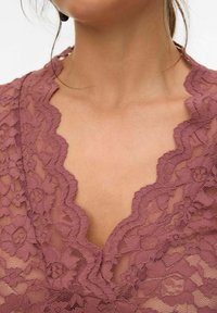 Vêtement en dentelle mauve avec un décolleté en V profond, présentant des motifs floraux et des bords festonnés, mettant en valeur une texture et des détails complexes.