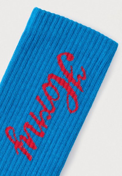 Μπλε κάλτσα με ριβ που διαθέτει κόκκινο κεντημένο κείμενο με παιχνιδιάρικη γραμματοσειρά. Το ύφασμα έχει υφή πλεκτού με ανάγλυφη εμφάνιση.