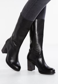 Bottes hautes en cuir noir à la texture lisse, bout arrondi et talon en bois épais. Présentent des coutures minimales et une fermeture éclair latérale.