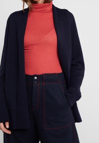 Cardigan azul marino con apertura frontal, llevado sobre un cuello alto rojo. Pantalones oscuros con costuras rojas y dos bolsillos frontales con botones.