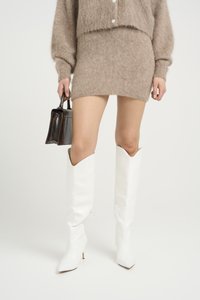 Hvite knelang støvler med spiss tå og gullhelle, laget av glatt materiale. Kombinert med en beige, lodden genser og miniskjørt.