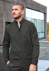 Camisa de botones de tela gris oscuro con un patrón geométrico texturizado, mangas largas y un cuello casual. Combinada con pantalones oscuros.