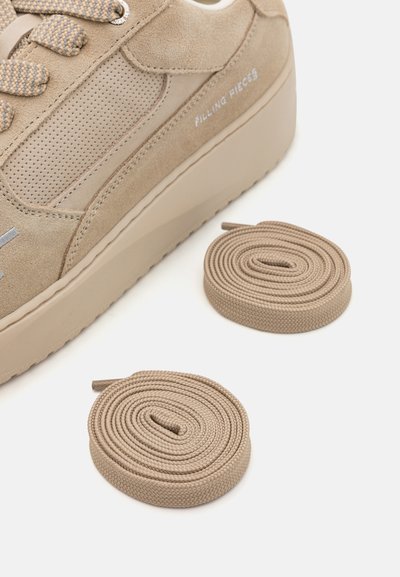 Zapatillas de ante beige con detalles perforados y acentos grises, acompañadas de dos cordones planos a juego sobre un fondo blanco.