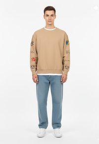 Svetter beige con stampe grafiche sulle maniche, colletto rotondo sopra una maglietta bianca, abbinato a pantaloni azzurri chiari in tessuto testurizzato e sneakers bianche.