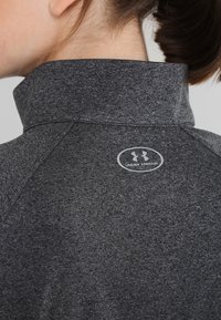 Top sportivo grigio con colletto rialzato. Presenta un tessuto testurizzato e un piccolo logo circolare di Under Armour sul retro.