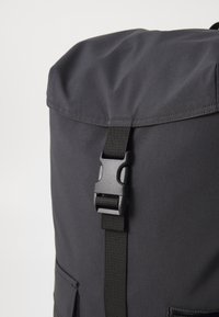 Black fabric backpack with a roll-top closure and a buckle strap. Features a textured surface and a front pocket with a flap.