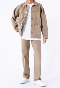 Chaqueta de mezclilla beige con grandes bolsillos y cierre de botones, combinada con pantalones beige a juego y zapatillas blancas. Diseño simple y casual.