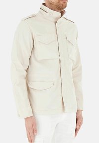 Giacca beige leggera con collo alto, quattro tasche frontali, polsini con bottoni e sottili dettagli di cucitura. Tessuto di consistenza liscia.