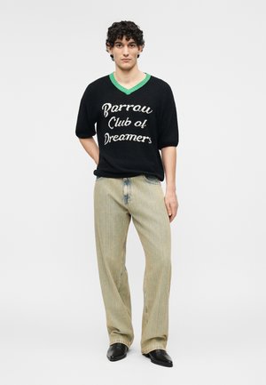 Joven con cabello rizado que lleva un suéter negro con ribete verde que dice "Barrow Club of Dreamers", jeans beige de pierna ancha y zapatos negros.