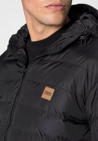 Doudoune noire avec fermeture éclair, surface texturée, poignets élastiques et écusson logo sur la poitrine. Conception à capuche.