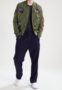 Giacca bomber verde oliva con vari patch, abbinata a pantaloni larghi blu navy e sneakers bianche. Camicia a righe visibile sotto.