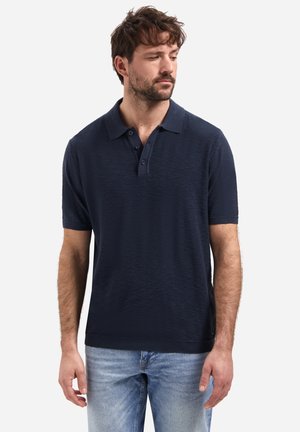 Man draagt een marineblauw poloshirt met korte mouwen en lichtblauwe spijkerbroek, kijkt naar beneden en naar links tegen een witte achtergrond.