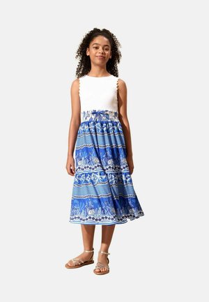 Mädchen mit lockigem Haar, das ein ärmelloses weißes Oberteil und einen blauen gemusterten Midirock mit Sandalen vor einem schlichten Hintergrund trägt.