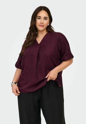 Donna con lunghi capelli castani che indossa una camicetta viola a maniche corte e pantaloni neri, posando davanti a uno sfondo chiaro e uniforme.