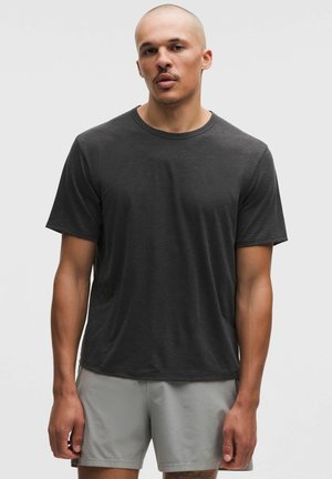 Man draagt een zwart T-shirt met korte mouwen en ronde hals en lichtgrijze korte broek, staande tegen een effen grijze achtergrond.