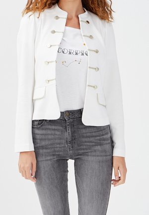 Femme portant une veste décorative blanche boutonnée sur un t-shirt blanc avec la constellation du Scorpion et un jean gris, visible des épaules jusqu'aux hanches.
