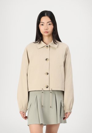 Beige cropped jacket with a collared neckline, large front buttons, elastic cuffs, and adjustable drawstring hem. Styled with a green pleated skirt.