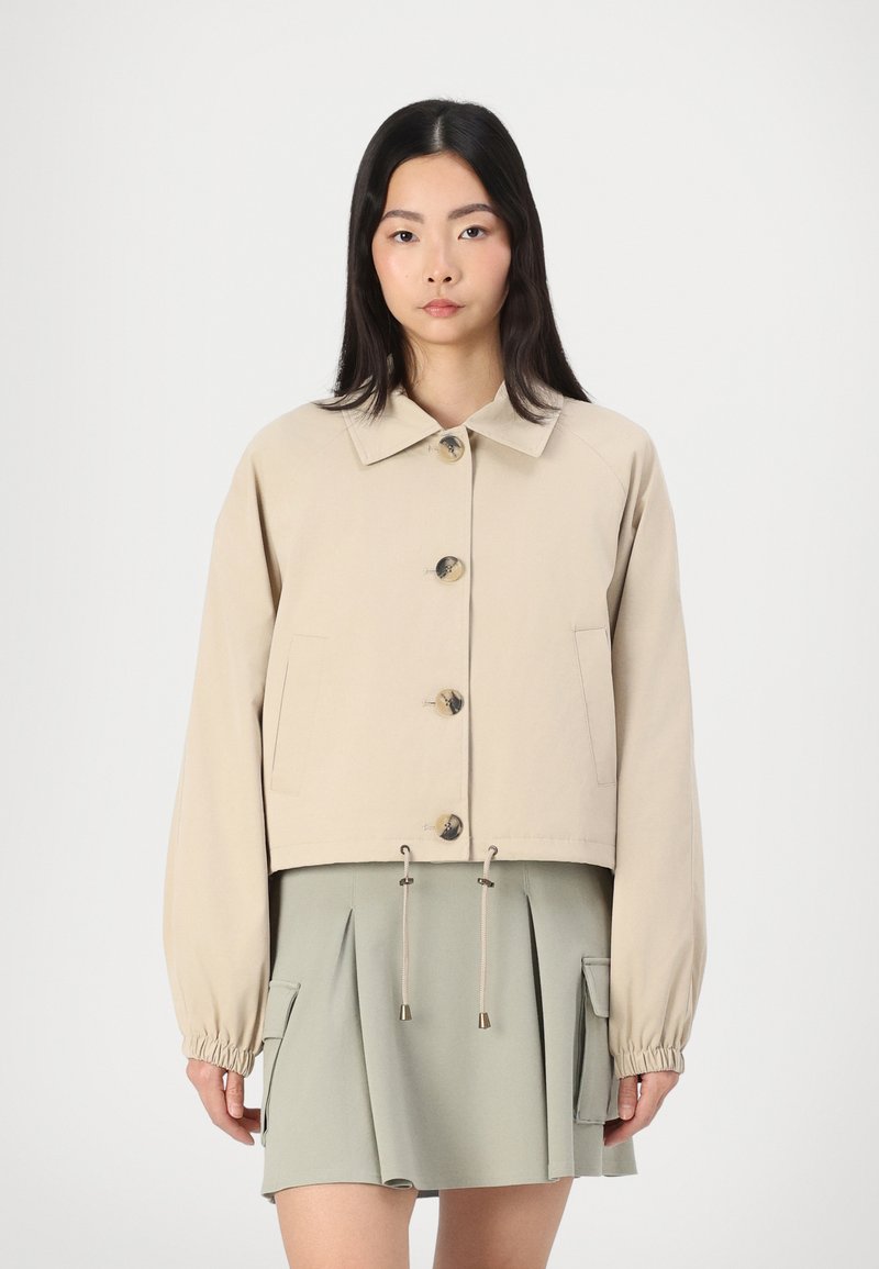Beige cropped jack met een kraag, grote knopen aan de voorkant, elastische manchetten en een verstelbare trekkoordzoom. Gestyled met een groene geplooide rok.