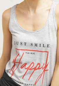 Šedý bavlněný tílko s kulatým výstřihem. Na přední straně je černý nápis "JUST SMILE" a "THINK HAPPY" uvnitř červeného obdélníkového rámu.