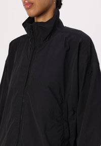 Giacca nera con zip caratterizzata da un colletto alto, texture leggera e branding discreto sulla manica. Tessuto liscio con linee di design pulite.