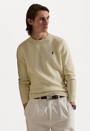 Lichtbeige gebreide trui met een geribde kraag en manchetten, voorzien van een klein zwart logo op de borst. Draag het met crèmekleurige broek.