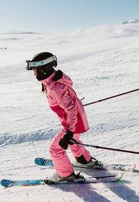 Růžový lyžařský oblek s kapucí a černými detaily, doplněný o černé rukavice. Lyže jsou modré s vzory a na hlavě je černá helma s brýlemi.