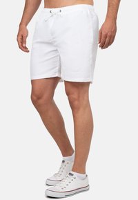 Pantaloni da bagno bianchi con cintura elastica e lacci, caratterizzati da una texture di tessuto liscia e tasche laterali. Indossati con sneakers bianche.