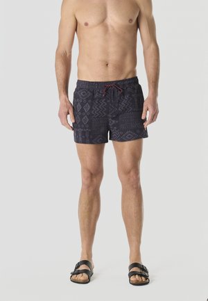 Torso y piernas masculinas vistiendo pantalones cortos de baño negros con estampado y cordón granate, y sandalias negras, de pie frente a un fondo gris liso.