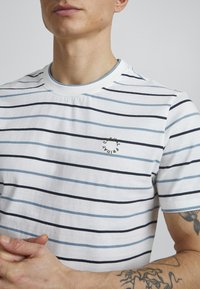 Hvit t-skjorte med korte ermer og horisontale blå og svarte striper. Har rund hals og liten brodert logo på brystet.