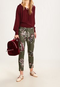 Burgundská blůza s vázáním kolem krku, kalhoty s olivovým květinovým vzorem a červený sametový batoh. Béžové šněrovací boty. Texturované látky s květinovými akcenty.