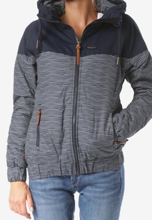 Kobieta ubrana w granatową kurtkę z kapturem z cienkimi białymi paskami, z przodu na zamek i z zamykanymi kieszeniami, w połączeniu z niebieskimi dżinsami.