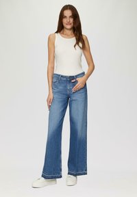 Weite blaue Jeans mit einem verblassten Waschung und gesticktem Saum, kombiniert mit einem eng anliegenden weißen Tanktop und weißen Turnschuhen.