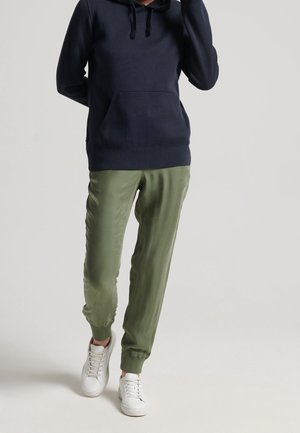 Persoon die olijfgroene joggerbroek, witte sneakers en een marineblauwe hoodie draagt, met gedeeltelijk zichtbare handen tegen een effen achtergrond.