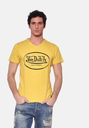 Homme portant un t-shirt jaune à col en V Von Dutch et un jean bleu délavé usé, debout les mains dans les poches sur un fond uni.