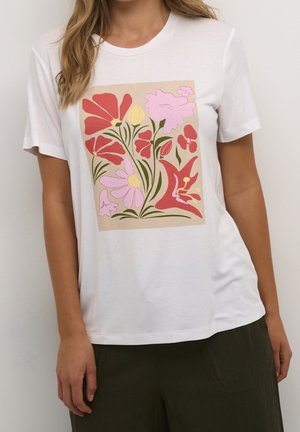 Nainen pukeutuneena valkoiseen t-paitaan, jossa on värikäs kukkakuosi, jossa esiintyy punaisia, vaaleanpunaisia ja keltaisia kukkia vihreine varrineen beigen taustan päällä.