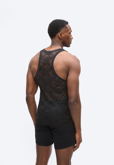 Άνδρας με κοντά μαλλιά που φοράει μαύρο αμάνικο γυμναστηρίου με σχέδιο και μαύρο σορτς, στέκεται με την πλάτη προς το μέρος μας, μπροστά σε ένα απλό φόντο.