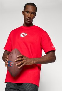 Sarkans kokvilnas T-krekls ar Kanzasitijas Čiefs logo uz krūšu, apvienots ar melniem šortiem. Personāžs tur brūnu NFL futbolu.