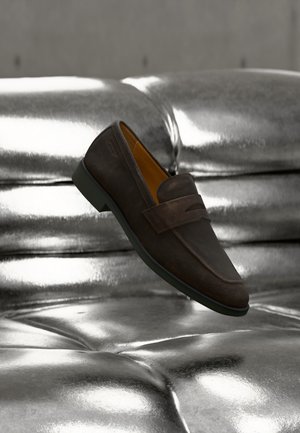 Brun læder loafersko med en lav sort sål, placeret mod en skinnende, tufted metallisk sølv pude baggrund.