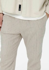 Pantaloni a righe beige con un tessuto texturizzato, caratterizzati da linee verticali in diverse sfumature. Include un piccolo etichetta nera sulla cintura.