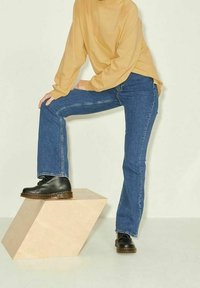 Jeans blu con taglio dritto, abbinati a un top lungo a maniche lunghe di colore giallo senape e stivali neri con lacci, posati su un piedistallo geometrico.