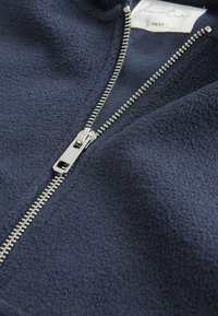 Marinblå fleece zip-jacka med silverfärgad metalldragkedja och mjukt material, med en etikett som säger "Danielle NEXT."