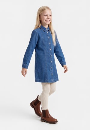 Ung pige smiler og går, iført en blå denimkjole med knapper, cremefarvede ribstrikkede leggings og brune ankelstøvler.