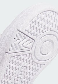 Bílá gumová podrážka tenisky s texturovanými vzory, včetně herringbone a kruhových designů, má hladký, mírně vyvýšený akcent.