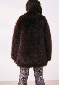 Manteau en fausse fourrure bordeaux au toucher doux et duveteux, avec un col haut et des manches longues. Porté avec des leggings à motifs.