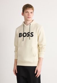 Jaunas vyras vilkintis béžinį džemperį su juodu "BOSS" užrašu, juodomis raištelėmis ir juodomis kelnėmis, stovintis prieš paprastą baltą fono.