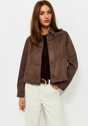 Vrouw met lang bruin haar die een bruine jas met knopen draagt, een zwart topje, een witte broek en een zwarte riem met een gouden gesp.