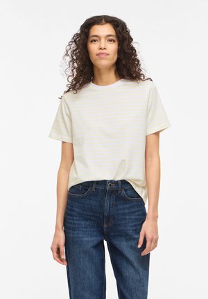 Mujer con cabello rizado, vistiendo una camiseta de rayas amarillas pálidas de manga corta y jeans azul oscuro, de pie frente a un fondo blanco.