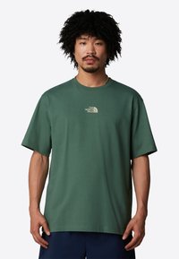 Camiseta de algodón verde con cuello redondo, mangas cortas y un pequeño logotipo beige en el pecho. Ajuste holgado con una textura suave.